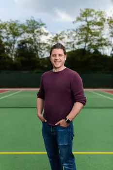 James, male sports engineer, smiles at camera on a tennis court