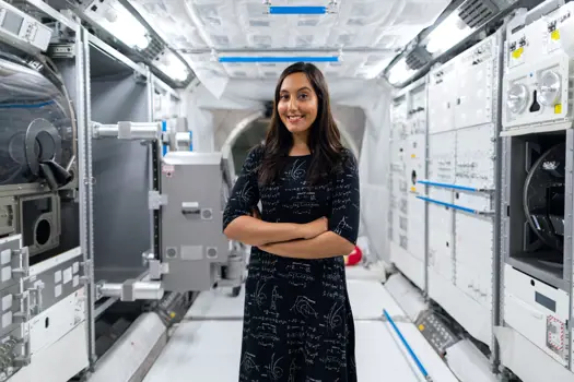 Vinita Marwaha Madill standing and smiling with her arms crossed with a background of space technology equipment.